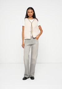 Gilet en polaire beige avec des boutons ronds et une bordure olive, associé à un t-shirt blanc et un jean évasé gris clair. Chaussures noires.