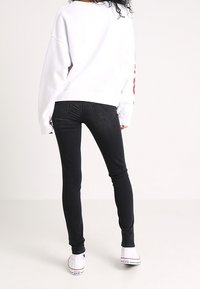 Persona con jeans negros ajustados, camisa blanca de manga larga y zapatillas altas blancas de pie sobre un fondo claro y sencillo.