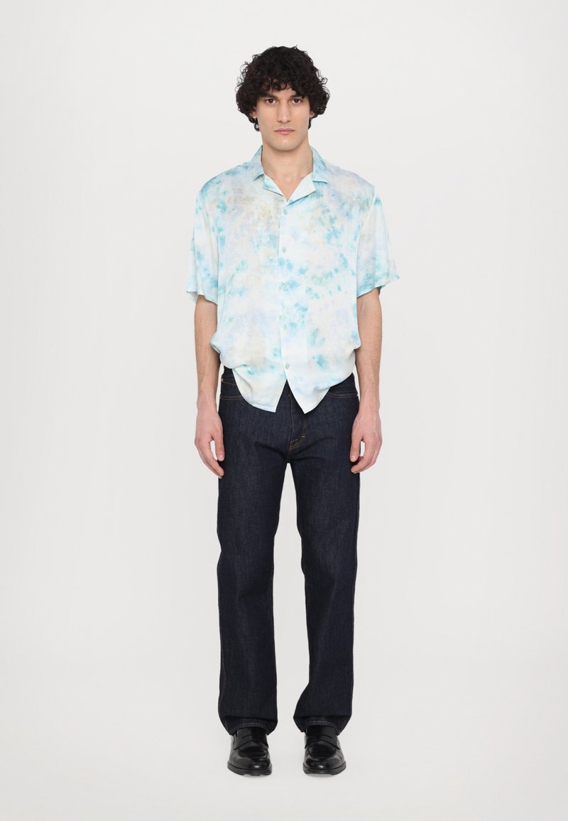 Kortærmet, knap-op skjorte med et lyseblåt tie-dye mønster, parret med mørkeblå jeans og sorte sko. Ensfarvet hvid baggrund.