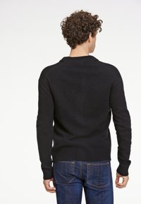 Svart stickad tröja med ribbad rund hals och mudd, med en slät yta och en figurnära design, i kombination med blå denimjeans.