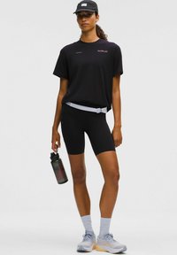 Czarny sportowy t-shirt, dopasowane czarne szorty, jasnoszare skarpetki za kostkę oraz szare buty do biegania z pomarańczowymi podeszwami. Trzyma ciemną butelkę na wodę.