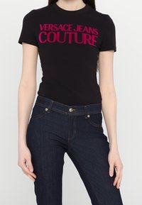 Svart bomullströja med stora rosa "VERSACE JEANS COUTURE" bokstäver, i kombination med mörkblå jeans i denim med gyllene detaljer och sömmar.