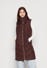 Gilet en duvet bordeaux avec un col montant, fermeture éclair dorée, motif matelassé en chevrons et poches latérales, porté sur un haut à manches longues blanc.
