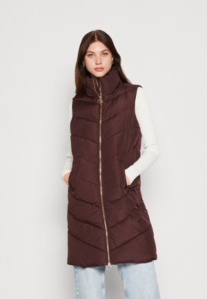 Gilet en duvet bordeaux avec un col montant, fermeture éclair dorée, motif matelassé en chevrons et poches latérales, porté sur un haut à manches longues blanc.
