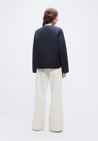 Γυναίκα με μακριά καστανά μαλλιά φοράει ένα ναυτικό μπλε καπιτονέ μπουφάν και λευκά παντελόνια με φαρδιά γραμμή, στέκεται με την πλάτη προς το μέρος μας μπροστά σε ένα απλό φόντο.