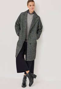 Manteau gris texturé avec un large revers, porté sur un pull gris clair, associé à un pantalon noir à jambes larges et des bottines noires.