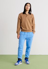 Bruine lange mouw polo met zwarte kraag, gecombineerd met blauwe trainingsbroek en blauwe Nike sneakers. Eenvoudig ontwerp, zachte textuur.