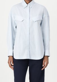 Ljusblå och vit randig knapplik skjorta med spetsig krage och bålfickor, bärs tillsammans med mörkblå byxor av en stående person.