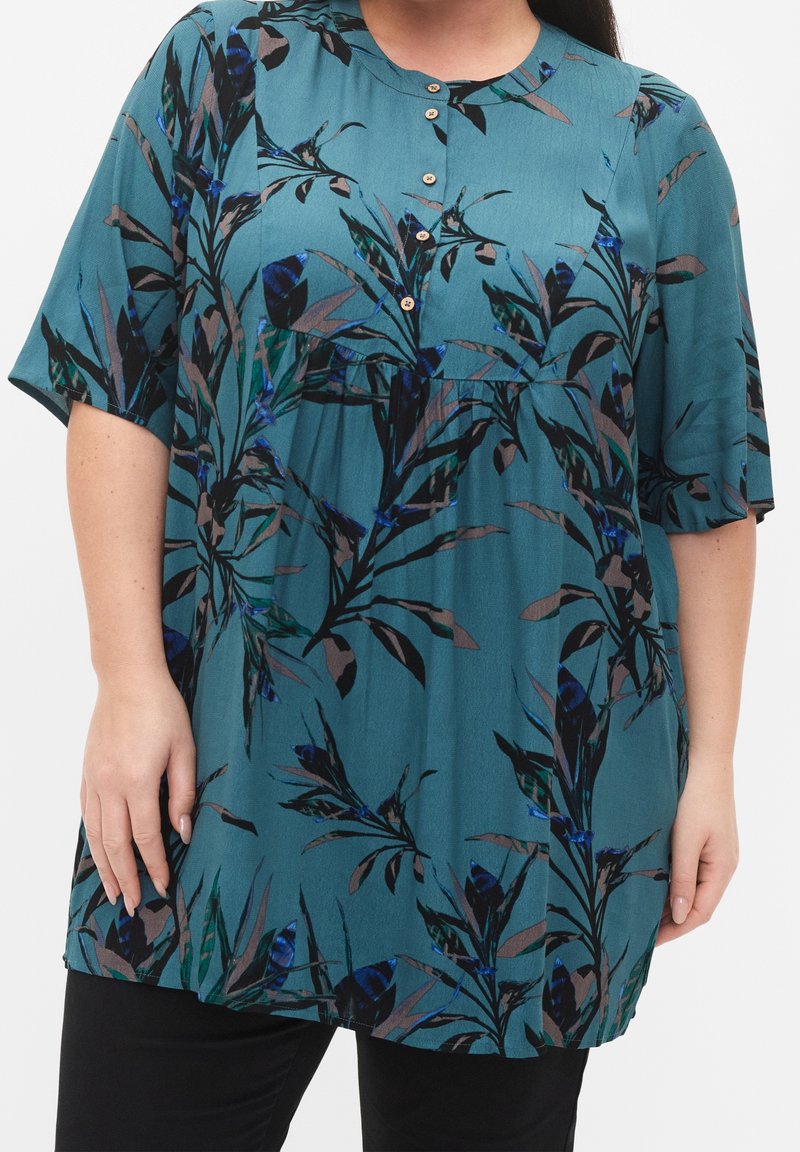 Blouse à manches courtes couleur sarcelle avec imprimé botanique noir, bleu et marron et petits boutons sur le devant, portée avec un pantalon noir.