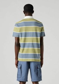 Polo a maniche corte in verde, blu e strisce bianche. Collo bianco ben evidente. Abbinato a pantaloni cargo blu con tasche laterali.