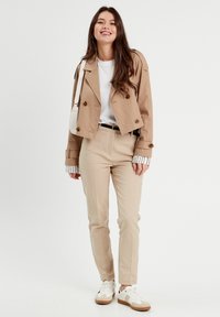 Beige cropped jacket with brown buttons, paired with beige trousers and white shirt. White sneakers with brown accents. Striped sleeve detail.