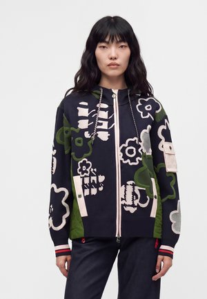 Femme aux longs cheveux noirs portant un sweat à capuche zippé sombre avec de grands motifs floraux abstraits en vert, blanc et rose, debout devant un fond uni.