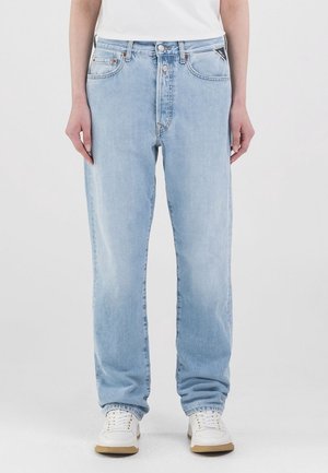 Personne portant un jean ample bleu clair, des baskets blanches et une chemise blanche, debout devant un fond clair uni.