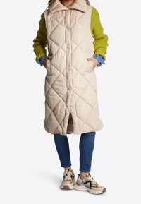 Gepolsterte lange Weste in Hellbeige mit großem Kragen, seitlichen Taschen und Knopfverschlüssen, getragen über einem grünen Strickpullover und blauen Jeans.