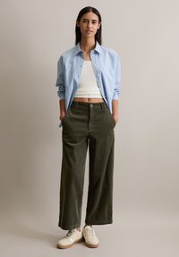 Hellblau gestreiftes Button-Up-Hemd über einem weißen gerippten Tanktop, kombiniert mit dunkelgrünen, weiten Cordhosen und weißen Turnschuhen.