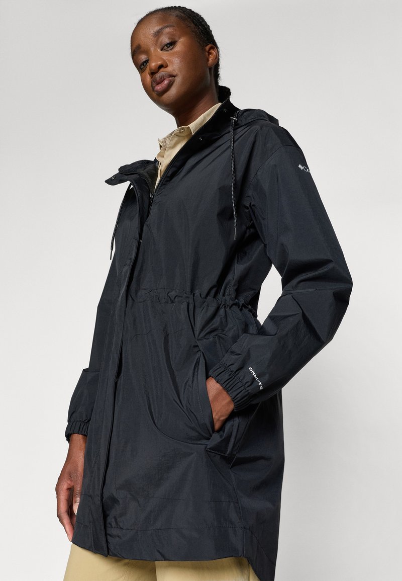 Veste noire légère avec une taille élastique, fermeture éclair à l'avant, poches et capuche ajustable. Présente des poignets froncés pour un ajustement confortable.