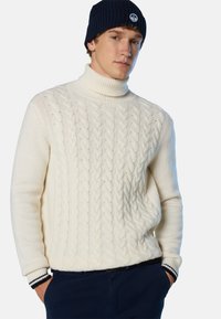 Cremfarbener Strickpullover mit Rollkragen und gerippten Bündchen, verziert mit navyblauen Streifen, kombiniert mit einer dunkelblauen Mütze mit einem Logo-Patch.