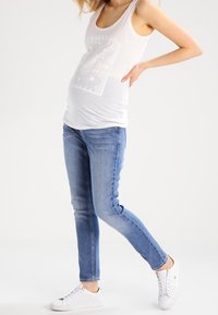 Bílé tílko s krajkovým vzorem, světle modré skinny džíny a bílé tenisky s perforovanými detaily, nošené ve stojící pozici.