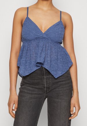 Mujer que lleva una blusa azul sin mangas con textura de ojales y dobladillo acampanado y vaqueros negros de cintura alta sobre un fondo liso.
