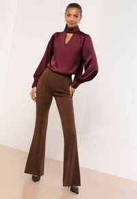 Burgunderfarbene Satinbluse mit einem Ausschnitt in Tropfenform, kombiniert mit braunen, eng anliegenden, ausgestellten Hosen. Das Outfit zeichnet sich durch lange Ärmel und glatte Texturen aus.