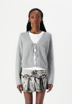 Light gray cropped cardigan with a V-neck and tie front, layered over a white ribbed top, paired with a silver-accented pleated skirt.