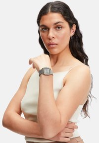 Orologio metallico quadrato color argento con cinturino a maglia, indossato su un polso. L'orologio presenta un semplice quadrante grigio e un design minimalista.