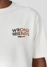 Krémové tričko s vyšitým logem "WRONG FRIENDS DESIGN HOUSE" v hnědé a černé barvě. Měkký materiál s krátkými rukávy.