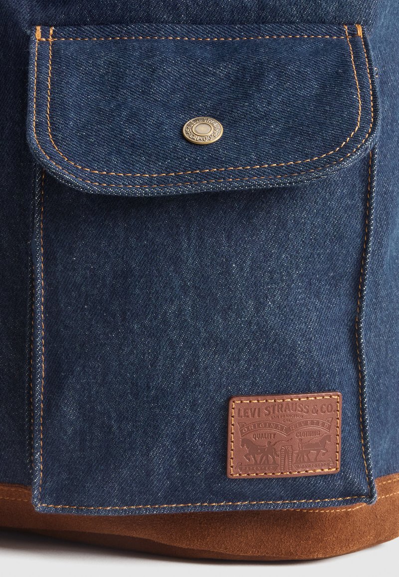 Borsa in denim con una finitura blu scuro, dotata di una tasca con chiusura a scatto, cuciture arancioni e un'etichetta in pelle marrone embossata con i dettagli del marchio.
