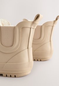 Paire de bottines de pluie beige à la cheville avec semelles texturées et languettes, présentant des panneaux latéraux élastiques et une doublure intérieure douce.