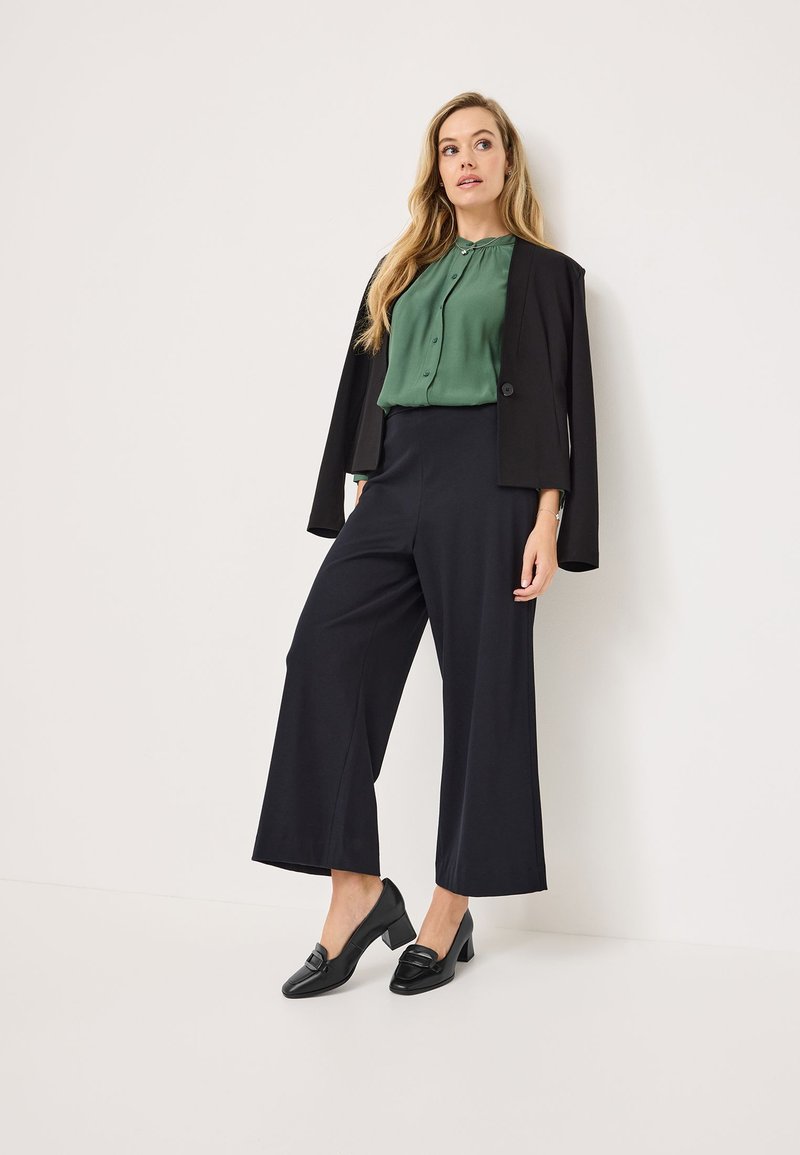Blazer noir sur une chemise verte à boutons, associé à un pantalon noir taille haute à jambes larges et des chaussures à talons noirs avec un détail à boucle.