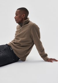 Lysbrun fleecepullover med lynlås til kvart hals, struktureret overflade og lange ærmer, parret med mørke jeans.