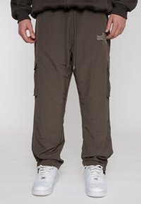 Cargo-broek in een lichtbruine, gestructureerde stof met zijzakken en een trekkoord in de taille, gedragen met witte sneakers.