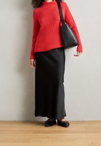 Rode geribbelde trui, zwarte aansluitende rok, zwarte schoudertas en zwarte platte schoenen met accentdetails. Tegen een neutrale achtergrond.