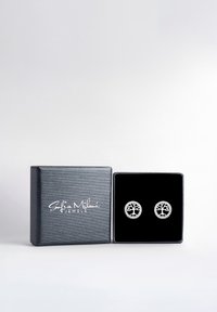 Silvriga örhängen med träddesign, placerade i en svart sammetdisplay inuti en texturerad svart smyckeslåda med vit logotyp.