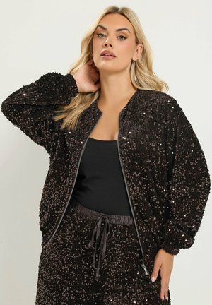Mujer rubia con chaqueta negra de lentejuelas con cremallera y pantalones de lentejuelas a juego con un top negro, posando con una mano detrás del cuello.