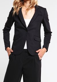 Blazer nero su misura con un design elegante, rever a punta e due tasche frontali. Tessuto liscio con una silhouette aderente.
