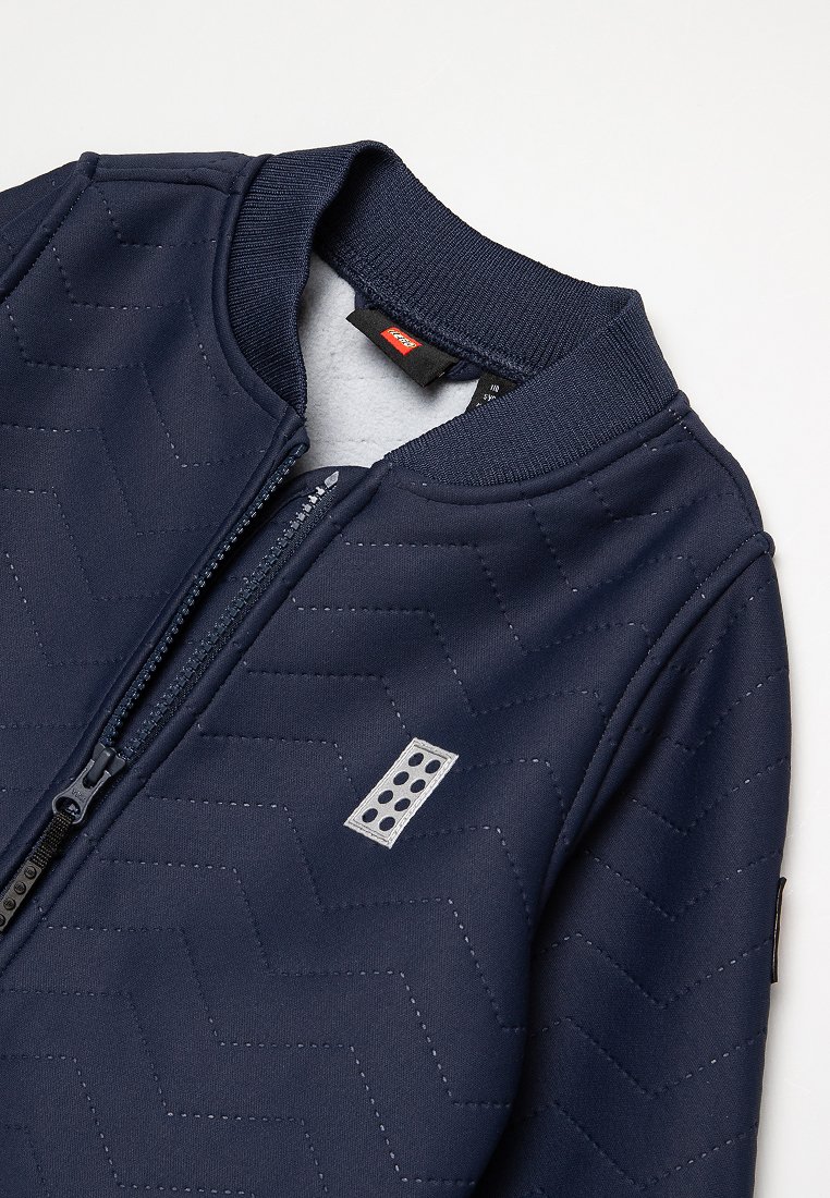 Giacca zip-up navy con pattern a rombi trapuntato, colletto e polsini a costine, e una toppa con logo che presenta un design stilizzato a mattoni.