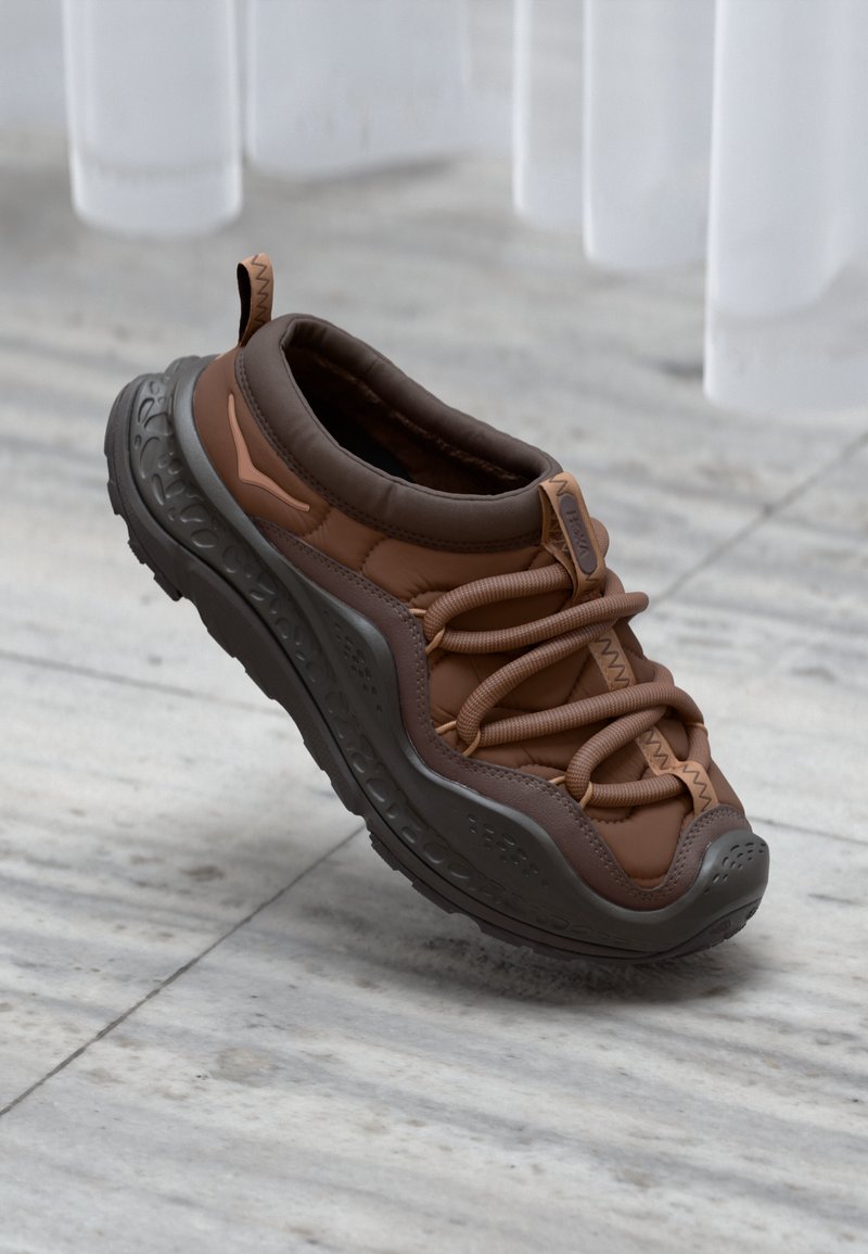 Zapatilla marrón con parte superior acolchada, cordones elásticos y suela de goma gris texturizada con un patrón elevado. Diseño ligero y sin ataduras.