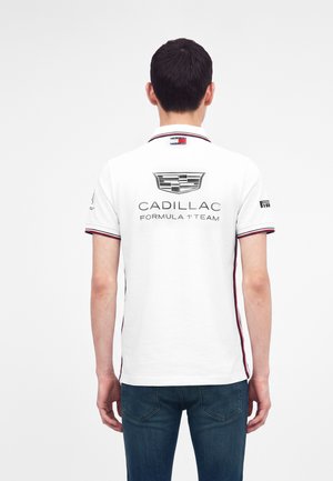 Mężczyzna ubrany w białą koszulkę polo z logo zespołu Cadillac Formula 1 na plecach i ciemnoniebieskie jeansy, stojący tyłem na jednolitym tle.