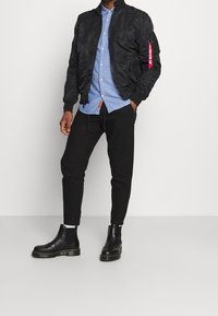 Giacca bomber nera con finitura testurizzata, camicia blu con bottoni, pantaloni neri a gamba affusolata e stivali Chelsea in pelle nera con suola robusta.