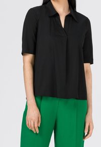 Blouse noire à manches courtes avec une coupe décontractée, col classique et patte de boutonnage à l'avant. Assortie à un pantalon vert vif pour un contraste.