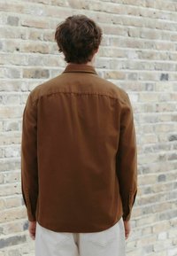 Hnedá košeľa na gombíky s dlhými rukávmi, s hladkou textúrou a zakriveným lemom. Zadný pohľad na svetlú tehlovú stenu.