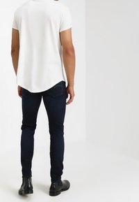 Vit kortärmad skjorta med rundad fåll, kombinerad med mörkblå slim fit-jeans och svarta ankelstövlar, sedd bakifrån.