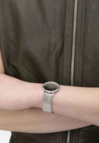 Rund svart klocka med silvernätband, bärs på ett handled. Bakgrunden visar en mörkgrön läderjacka med synlig dragkedjedetalj.