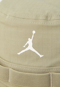Kaki spalvos kibiro skrybėlė su balta "Jumpman" logotipu. Strukturinė medžiaga su matomu tinkleliu, siuvinėti kraštai ir plati, plokščia kraštai.