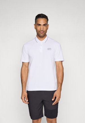 Polo bianco con colletto, tre bottoni e logo EA7. Presenta dettagli neri a contrasto sul colletto. Indossato con pantaloni corti neri.