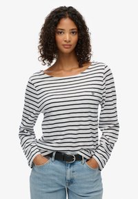 Gestreepte lange mouwen top in wit en marineblauw, met een wijde halslijn en geborduurd logo. Gecombineerd met lichtblauwe denim jeans en een riem.