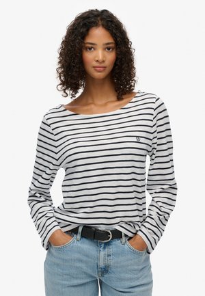 Maglietta a maniche lunghe a righe bianca e blu navy, con scollatura ampia e logo ricamato. Abbinata a jeans in denim blu chiaro e cintura.