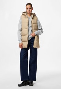 Beige Steppweste mit Kapuze, über einem grauen Sweatshirt getragen. Das Modell trägt zudem dunkelblaue weite Jeans und schwarze Stiefeletten.