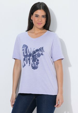 Kvinde iført lys lilla V-hals T-shirt med mørkeblå blomster-tekstgrafik, der siger "Flower", kombineret med mørkeblå jeans.